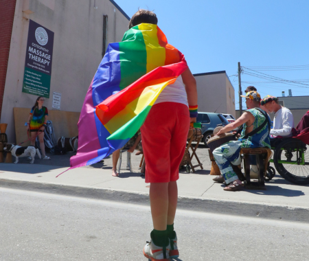 A young man on roller skates is wearing red shorts and a white shirt. He has a pride flag draped around his shoulders.